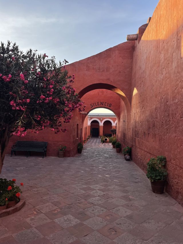 Cortile rosso terracotta con pavimentazione in pietra, albero in fiore rosa e un arco contrassegnato dalla parola SILENCIO.