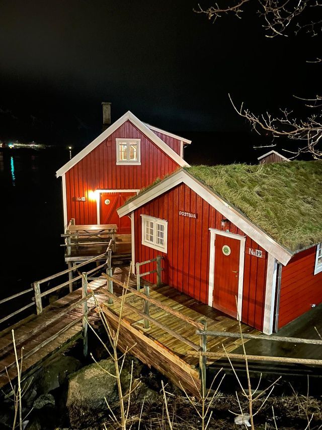 Due cabine in legno rosse e illuminate su palafitte sull'acqua di notte, una con tetto tradizionale in torba e collegate da una passerella.