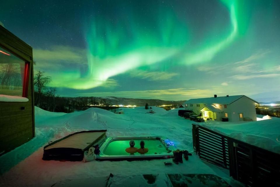 Due persone si rilassano in una vasca idromassaggio illuminata di notte, osservando l'aurora boreale in un paesaggio innevato.