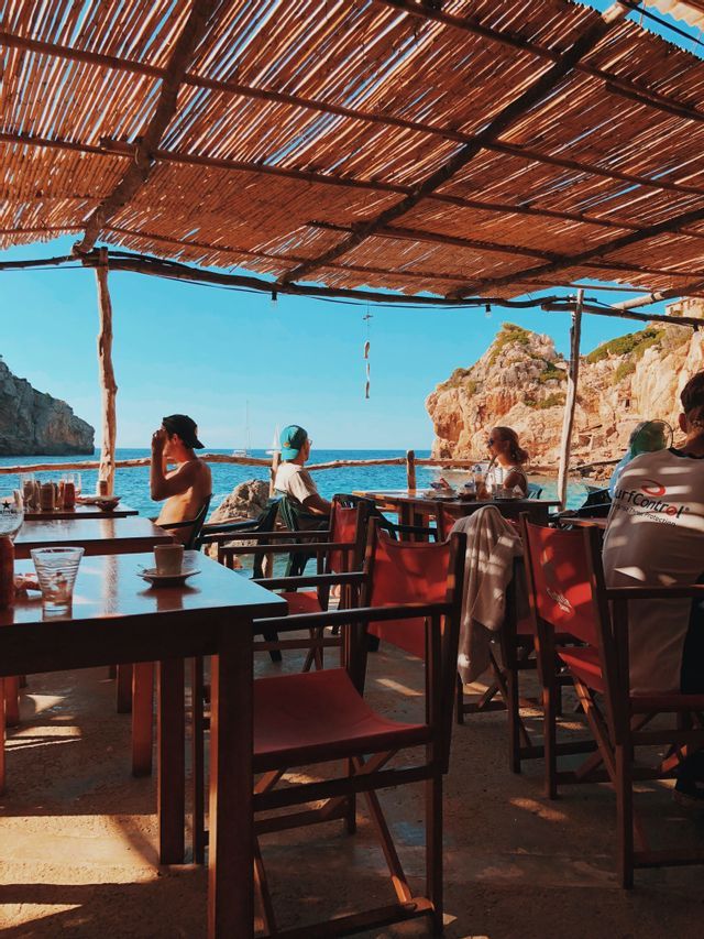 Un viaje en grupo de WeRoad relajándose en un café junto al mar bajo un techo de paja, con vistas al mar azul y acantilados rocosos.
