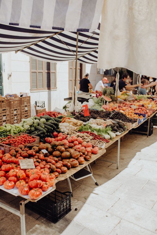 Eine Vielzahl von frischem Gemüse und Obst wird auf einem langen Tisch an einem Marktstand unter einem gestreiften Vordach präsentiert.