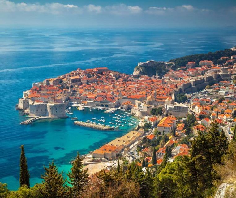 Una vista panorámica de una ciudad costera amurallada con tejados de terracota, rodeada por un mar azul en calma y un puerto lleno de barcos.