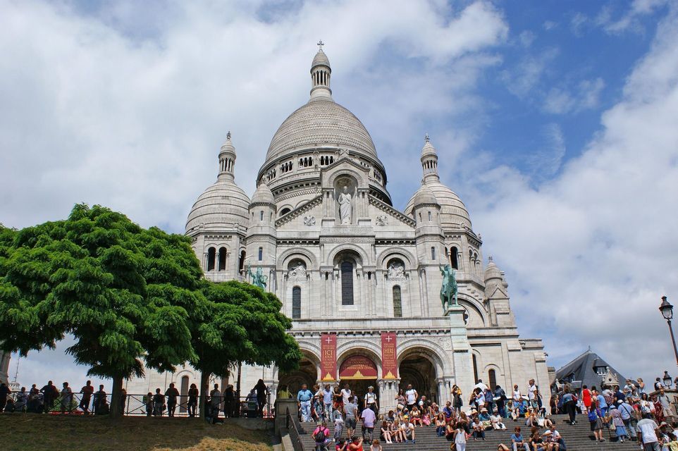 Folla di persone siede e cammina sui gradini all'esterno di una grande basilica con cupola bianca sotto un cielo parzialmente nuvoloso.