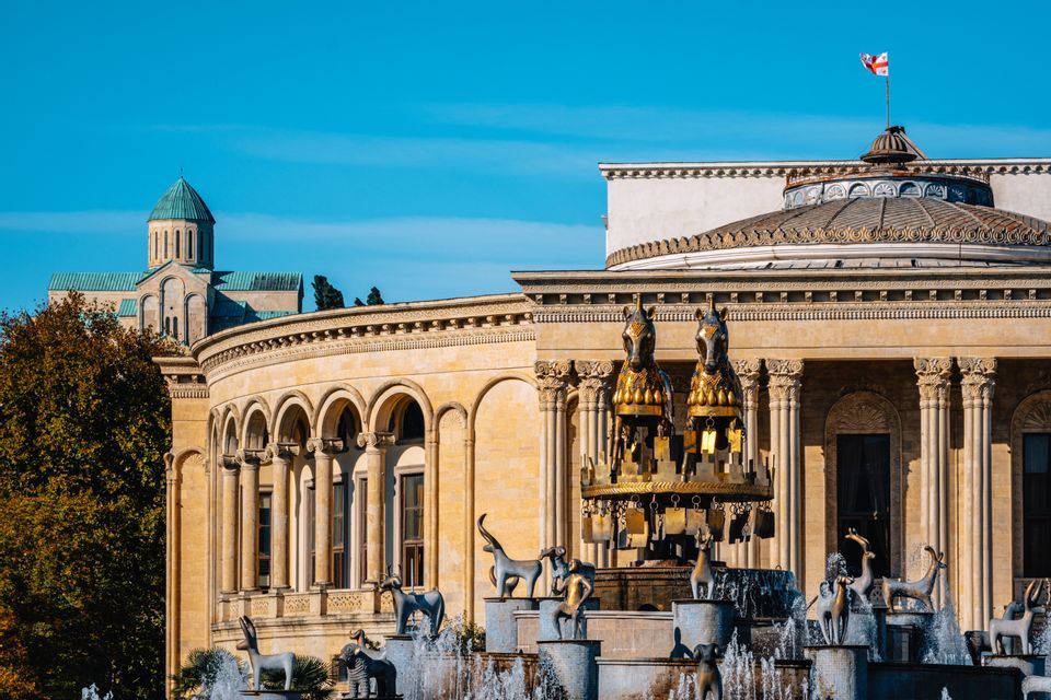 Una fuente de piedra con estatuas de animales y un caballo dorado se encuentra frente a un gran edificio con columnatas clásicas.