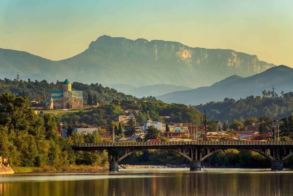 Un puente arqueado cruza un río tranquilo que conduce a un pueblo con una catedral en la colina frente a grandes montañas.