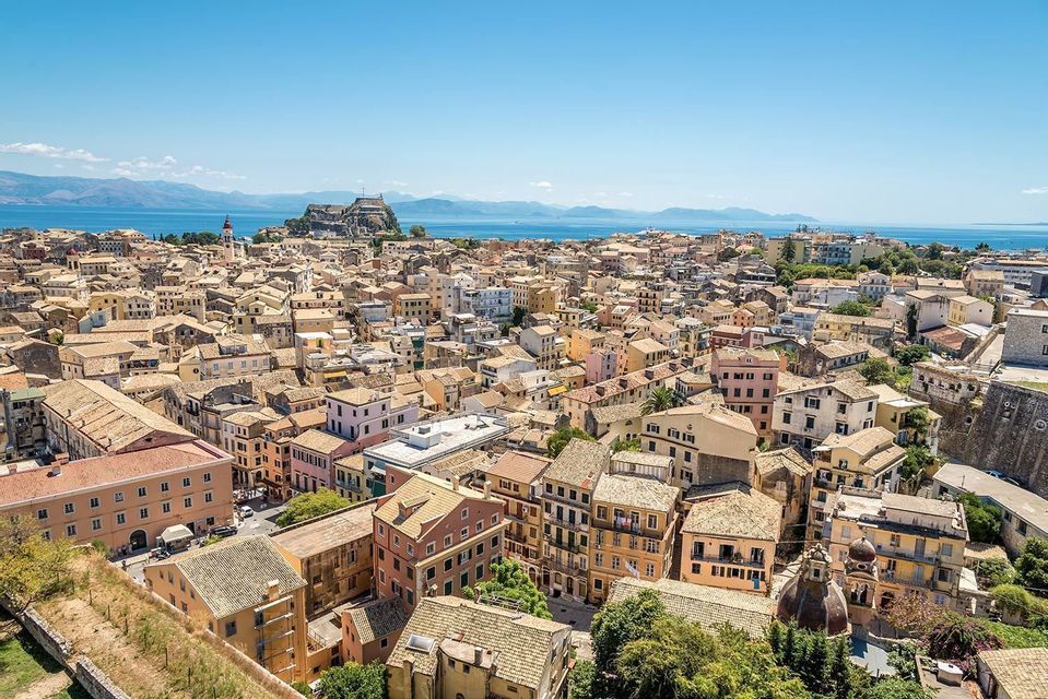 A high-angle view of a coastal city with terracotta roofs, with a fortress on the coast and mountains in the distance across the sea.