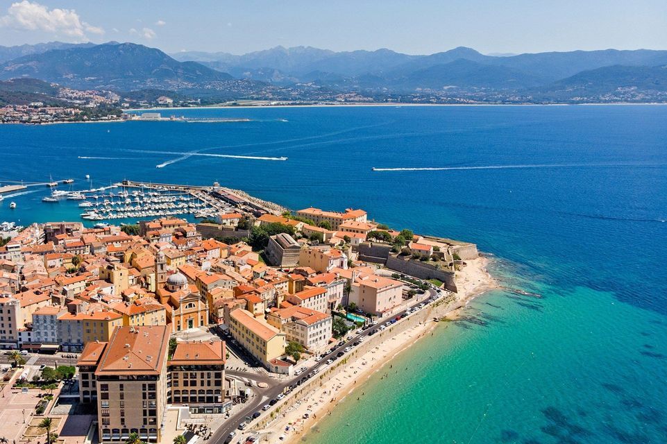 Vue aérienne d'une ville côtière avec un port, une plage de sable et une mer turquoise, avec des montagnes lointaines sous un ciel bleu.