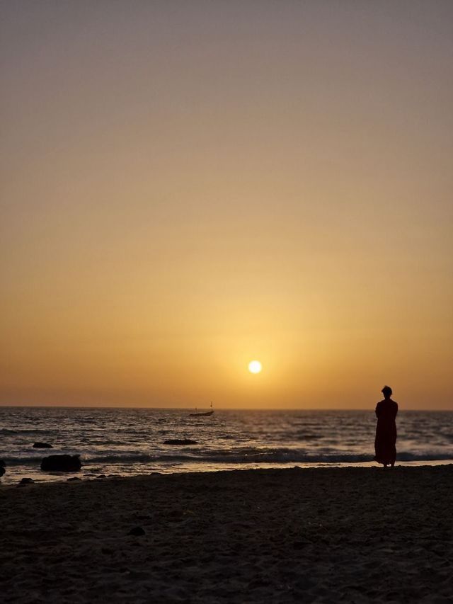 Una persona in silhouette si trova su una spiaggia sabbiosa a guardare il sole tramontare sull'oceano, con una piccola barca in lontananza.