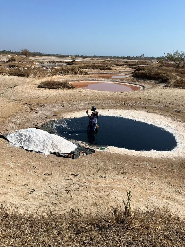 Una donna è immersa fino alla vita in una pozza d'acqua scura in una salina, con un grande mucchio di sale raccolto a terra accanto a lei.