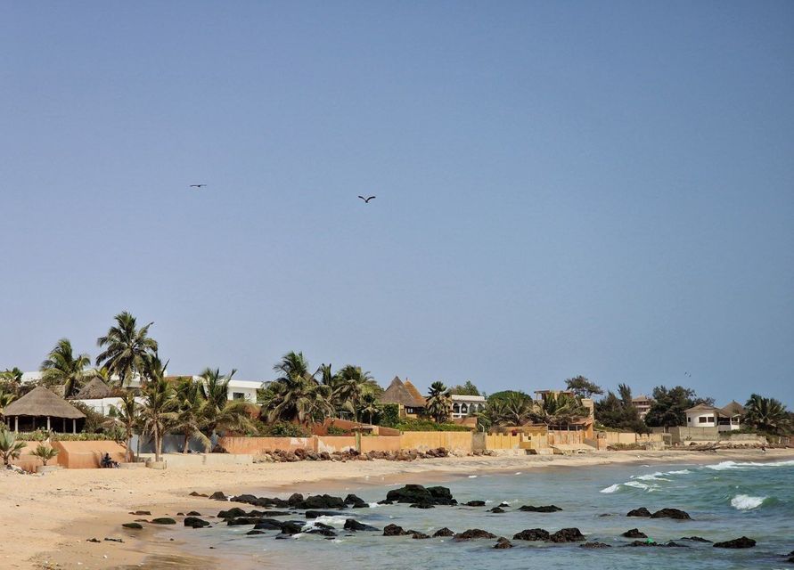 Un villaggio costiero con palme e edifici tradizionali si trova su una spiaggia sabbiosa accanto a una costa rocciosa sotto un cielo azzurro e limpido.