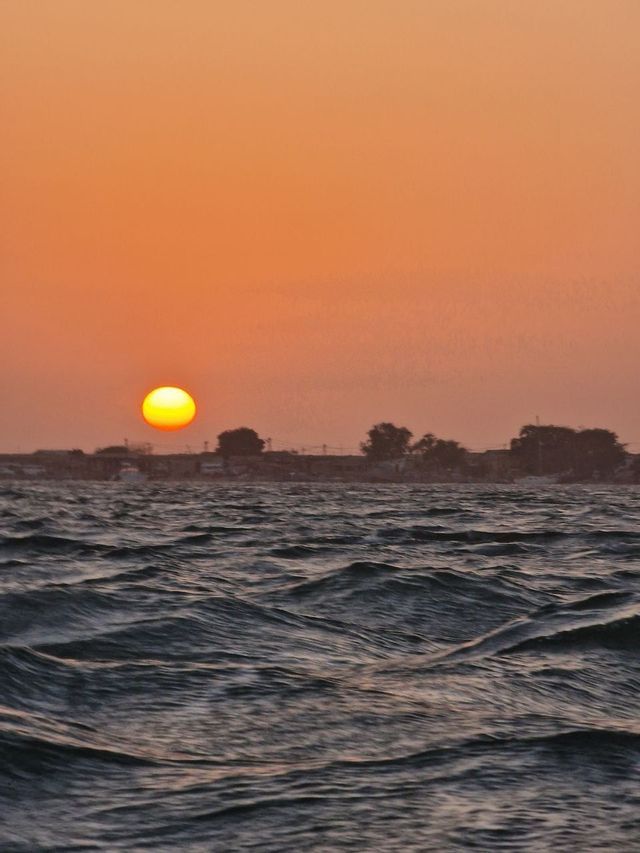 Il sole tramonta sul mare increspato, dipingendo il cielo di un bagliore arancione sopra una costa lontana.