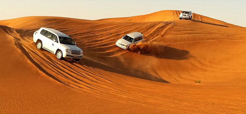 Three white SUVs drive across rolling orange sand dunes during a WeRoad group trip's desert safari.