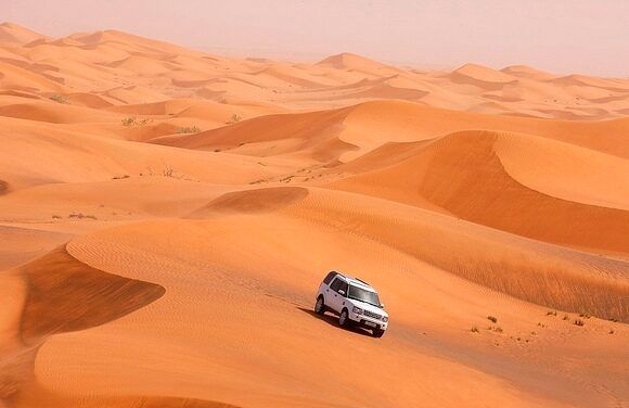 Un SUV bianco attraversa una vasta distesa di dune di sabbia ondulate e arancioni sotto un cielo velato.