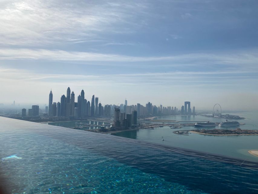 Una vista dal bordo di una piscina a sfioro che domina lo skyline di una moderna città costiera, con numerosi grattacieli e un porto.