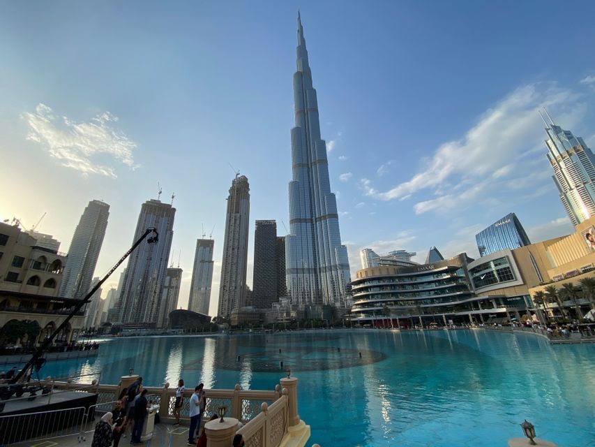 A towering skyscraper dominates a modern city skyline, viewed from across a large turquoise water feature under a clear sky.
