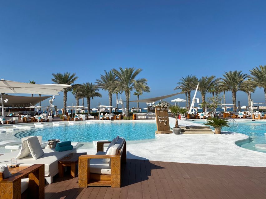 Un lussuoso resort balneare con una grande piscina circondata da lettini bianchi e palme sotto un cielo sereno.