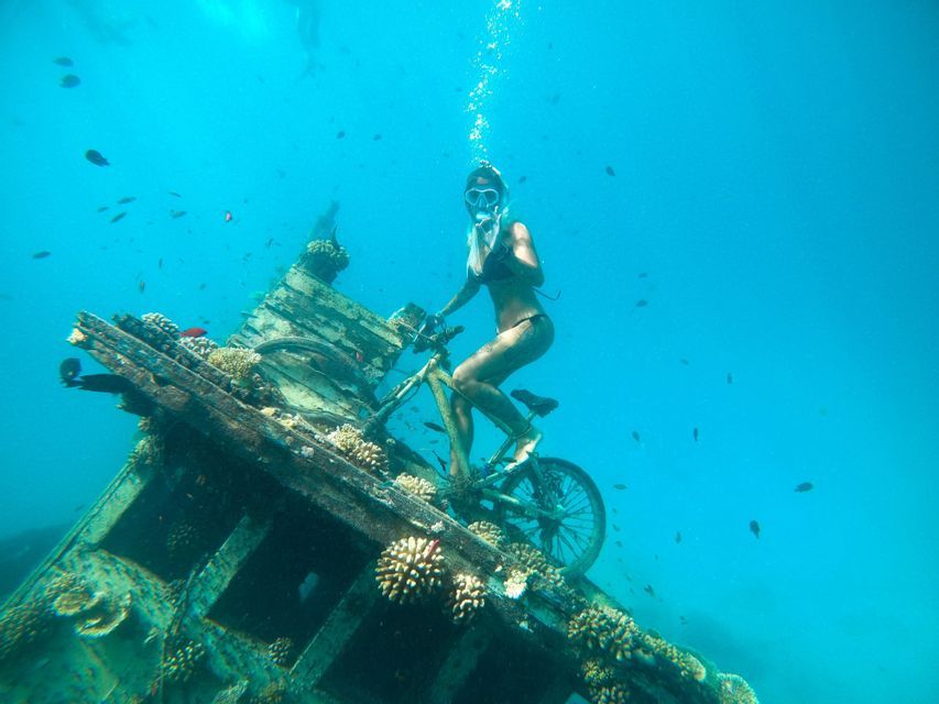 Una donna in bikini e maschera da sub siede in bicicletta su un relitto sommerso, circondata da acqua blu e piccoli pesci.