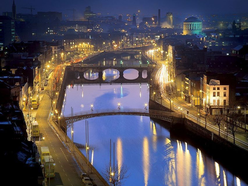 Una vista aerea di una città di notte, con un ampio fiume che la attraversa. Ponti, lampioni e scie luminose di auto si riflettono nell'acqua.