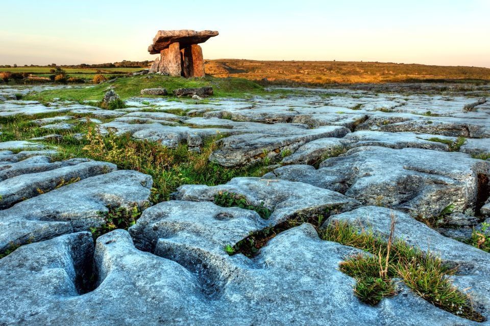 Un dolmen a portale preistorico si erge su un tumulo erboso in mezzo a una vasta pavimentazione calcarea fessurata al tramonto.