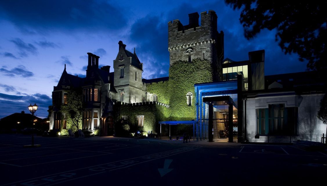 Un castello in pietra ricoperto d'edera con un ingresso moderno è illuminato da luci sotto un cielo notturno blu scuro.