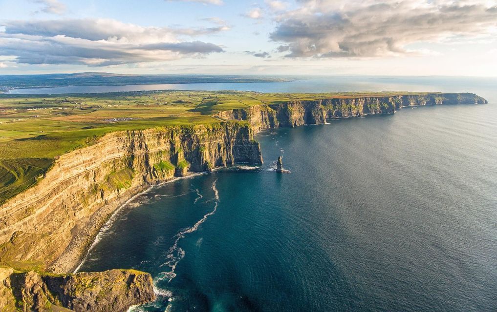 Una vista aerea di una costa spettacolare, dove scogliere a picco ed erbose incontrano il blu profondo dell'oceano sotto un cielo parzialmente nuvoloso.