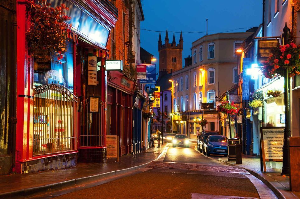 Una stretta strada di città bagnata è illuminata dalle luci dei negozi al crepuscolo, con edifici colorati e una torre di pietra in lontananza.