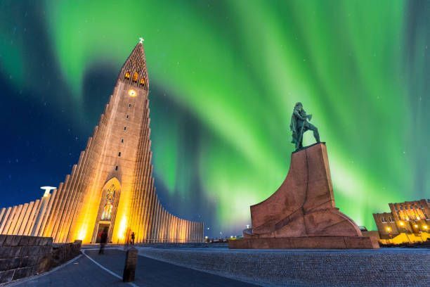 Una chiesa alta e moderna e una statua illuminata di notte, con l'aurora boreale verde che brilla nel cielo stellato.