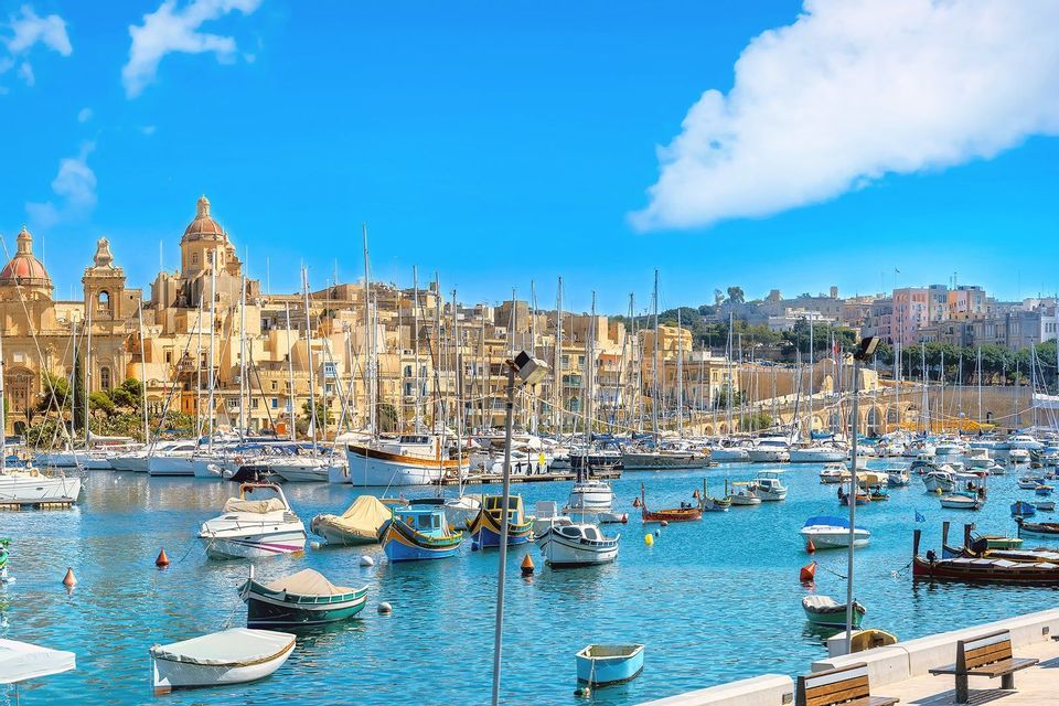 Ein geschäftiger Hafen voller Segelboote und kleiner Boote, mit einer historischen Stadtkulisse aus Steingebäuden unter einem strahlend blauen Himmel.