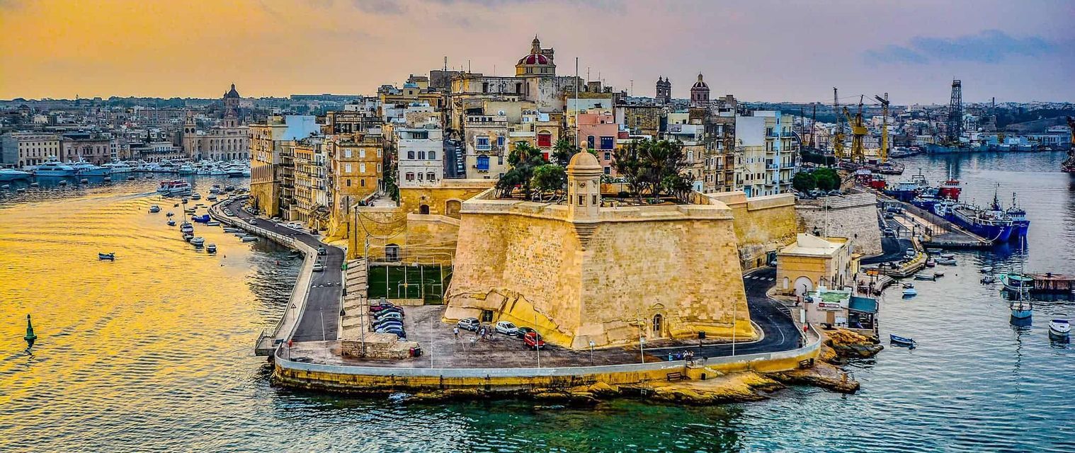 An aerial view of a fortified city on a peninsula jutting into a harbor filled with boats under a golden sunset sky.