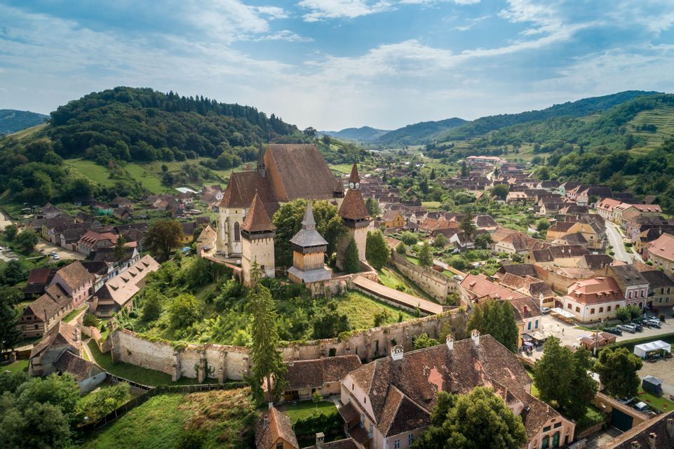 Eine Luftaufnahme einer historischen Wehrkirche auf einem Hügel, mit Blick auf ein kleines Dorf, eingebettet in ein grünes Tal.