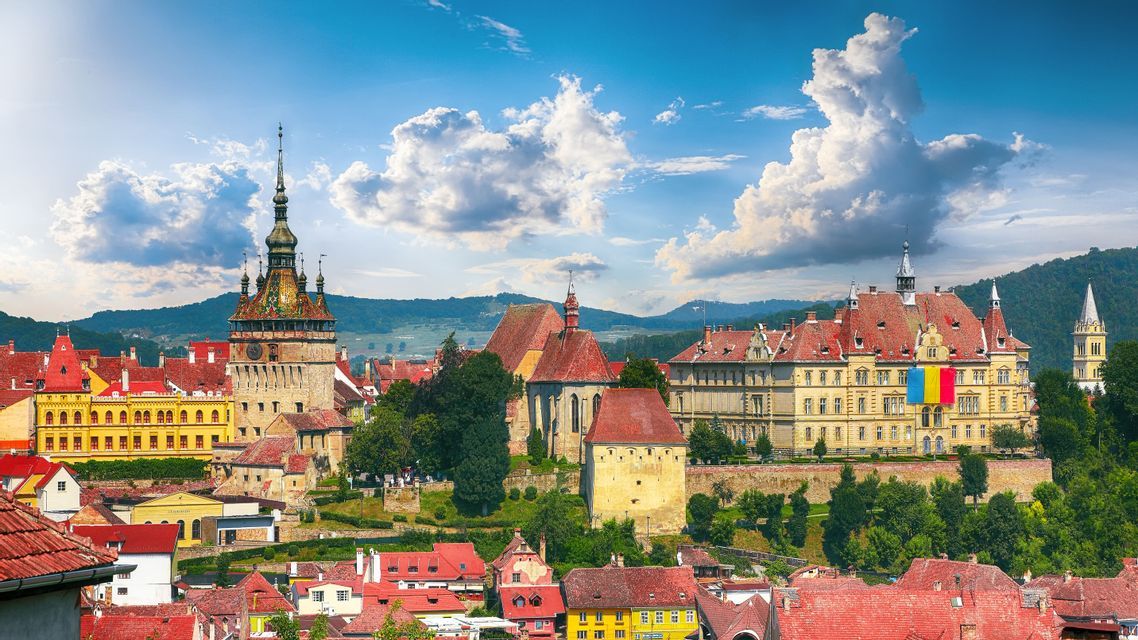 A historic town with a central clock tower and colorful buildings with red roofs, set against a backdrop of green hills and a blue sky.
