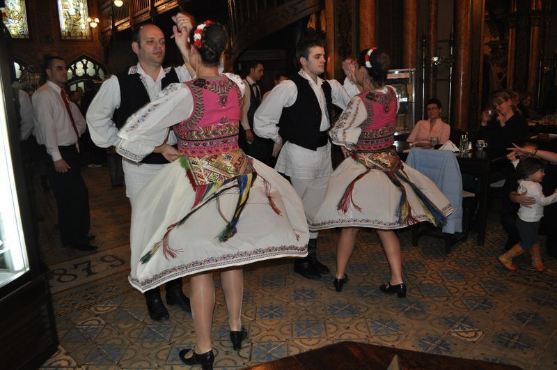 Two couples wearing colorful traditional costumes perform a folk dance for an audience inside a restaurant.