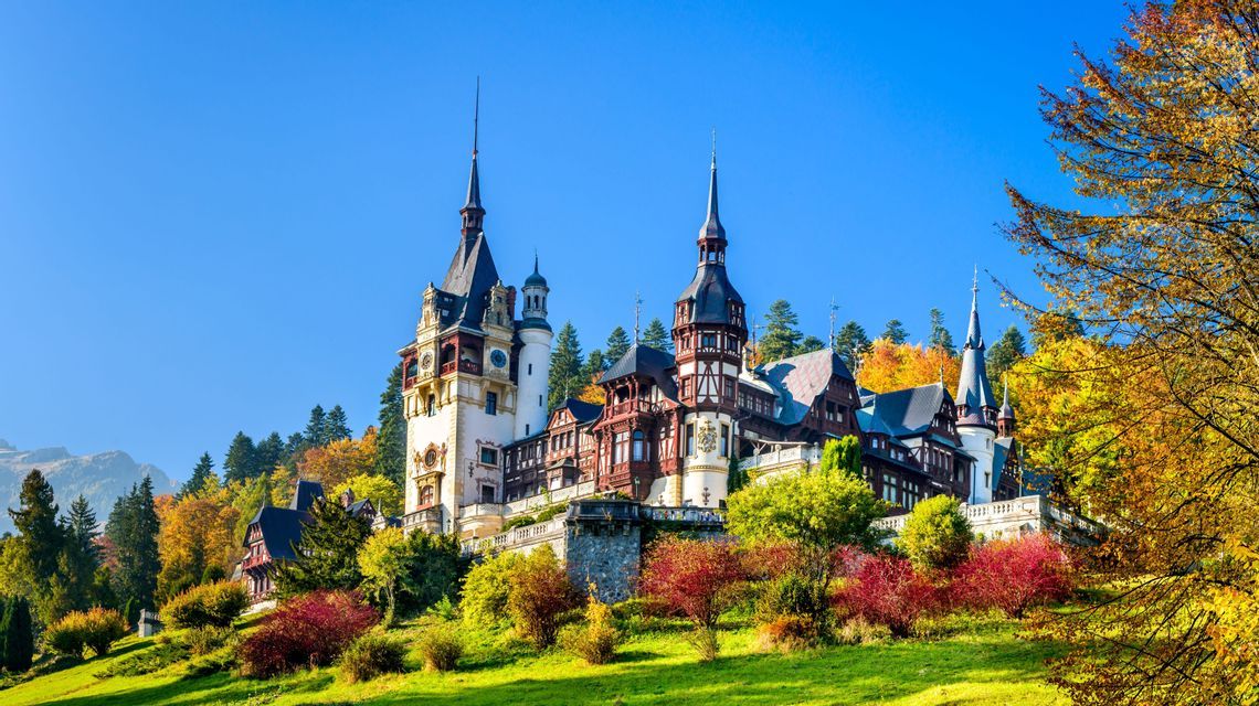 Ein reich verziertes Schloss mit mehreren Türmen thront auf einem grasbewachsenen Hügel, umgeben von buntem Herbstlaub vor einem klaren blauen Himmel.