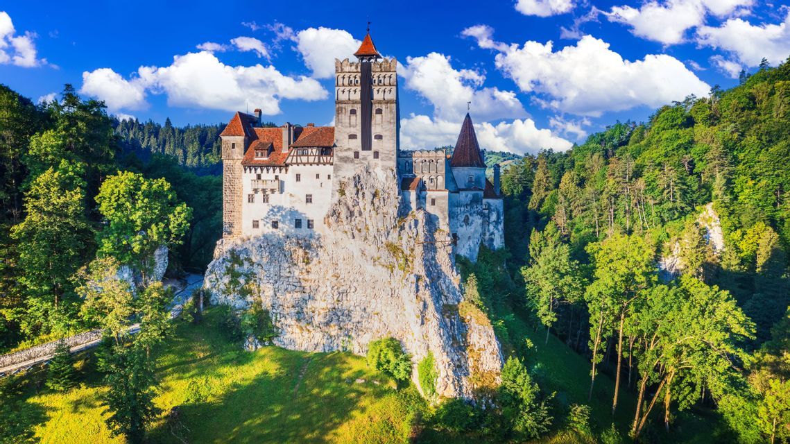 Ein steinernes Schloss mit roten Dächern steht auf einer felsigen Klippe, eingebettet in einen dichten grünen Wald unter einem teilweise bewölkten blauen Himmel.