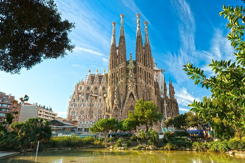 Una basilica riccamente decorata con molteplici guglie si erge sotto un cielo azzurro, vista da oltre un laghetto circondato da alberi rigogliosi.