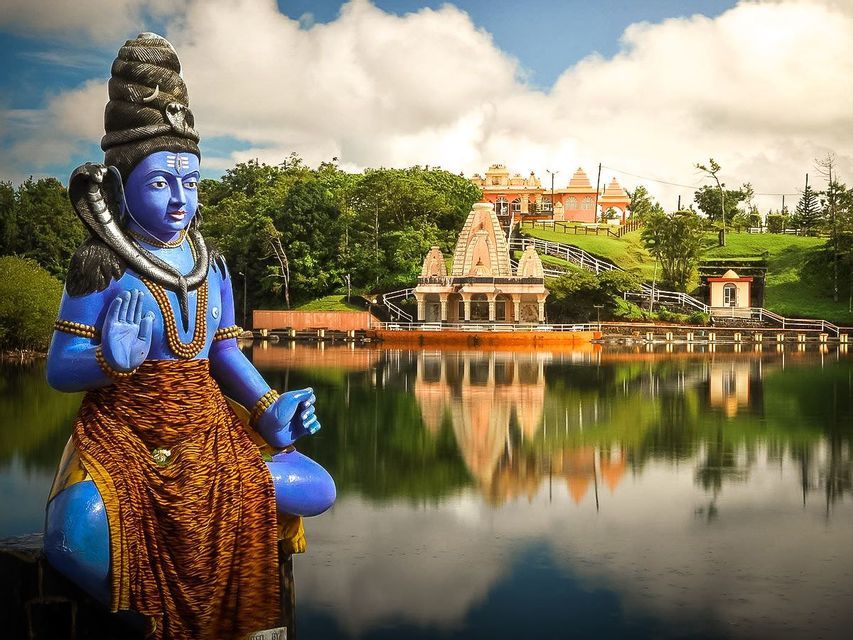 Una grande statua blu della divinità indù Shiva si trova vicino a un lago calmo, con un tempio sulla riva verde riflesso nell'acqua.