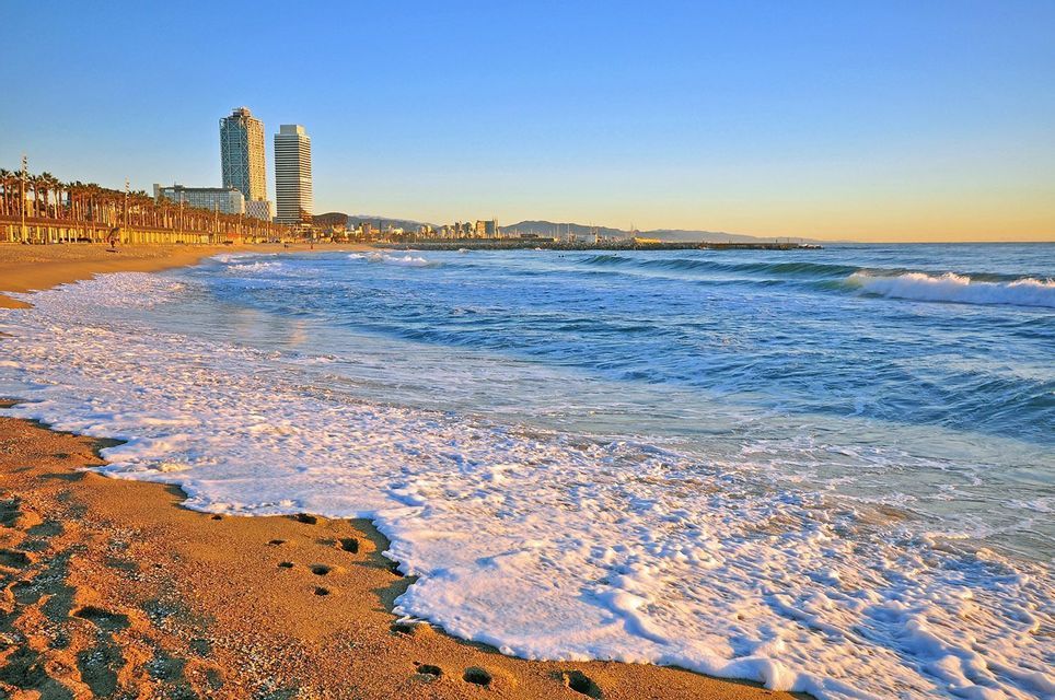 Onde oceaniche con schiuma bianca si infrangono su una spiaggia di sabbia dorata, con lo skyline di una città moderna in lontananza all'alba.