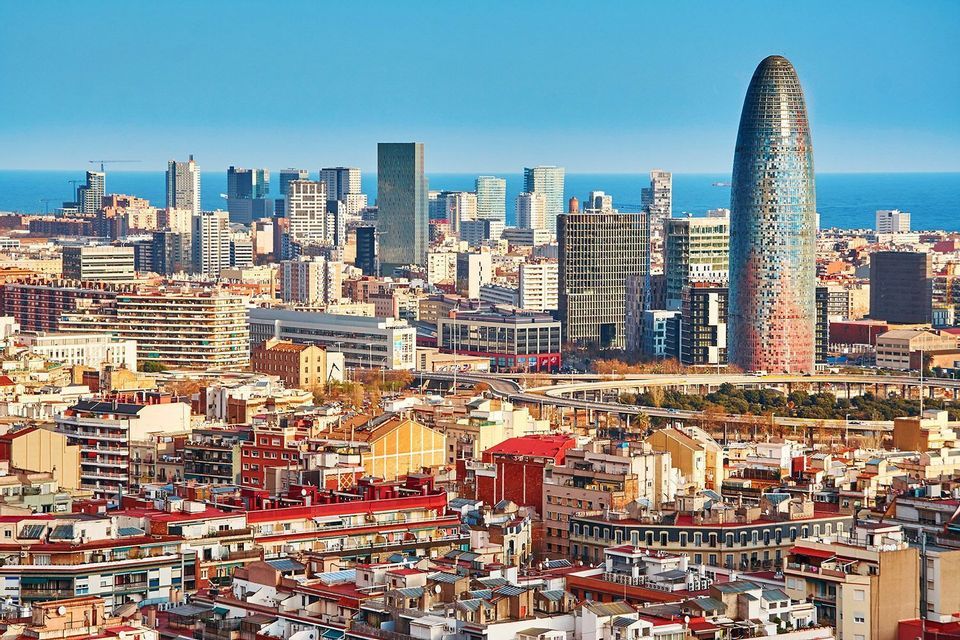 An expansive cityscape with diverse architecture, including a prominent bullet-shaped skyscraper, meets the sea under a clear blue sky.