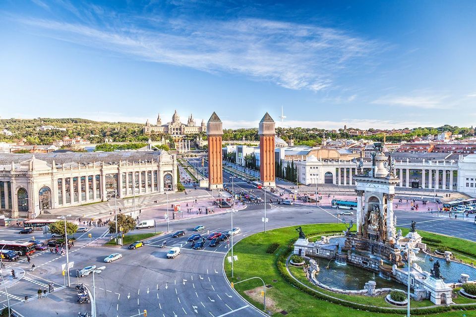 Una vista aerea di una grande piazza cittadina con una fontana decorativa, una rotonda e due torri di mattoni che conducono a un palazzo su una collina.