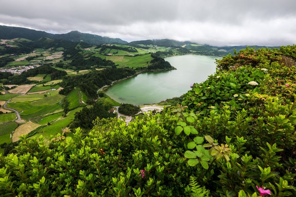 An aerial view of a green lake nestled in a valley of rolling hills and lush fields, under a cloudy sky.