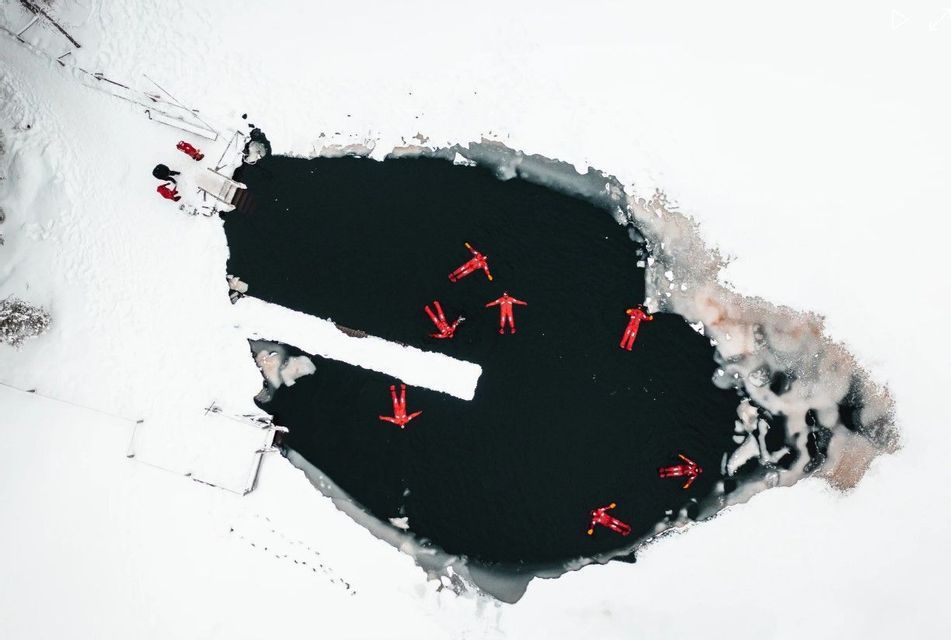 Veduta aerea di un viaggio di gruppo WeRoad con tute rosse che galleggiano in un'apertura in un lago ghiacciato e innevato.