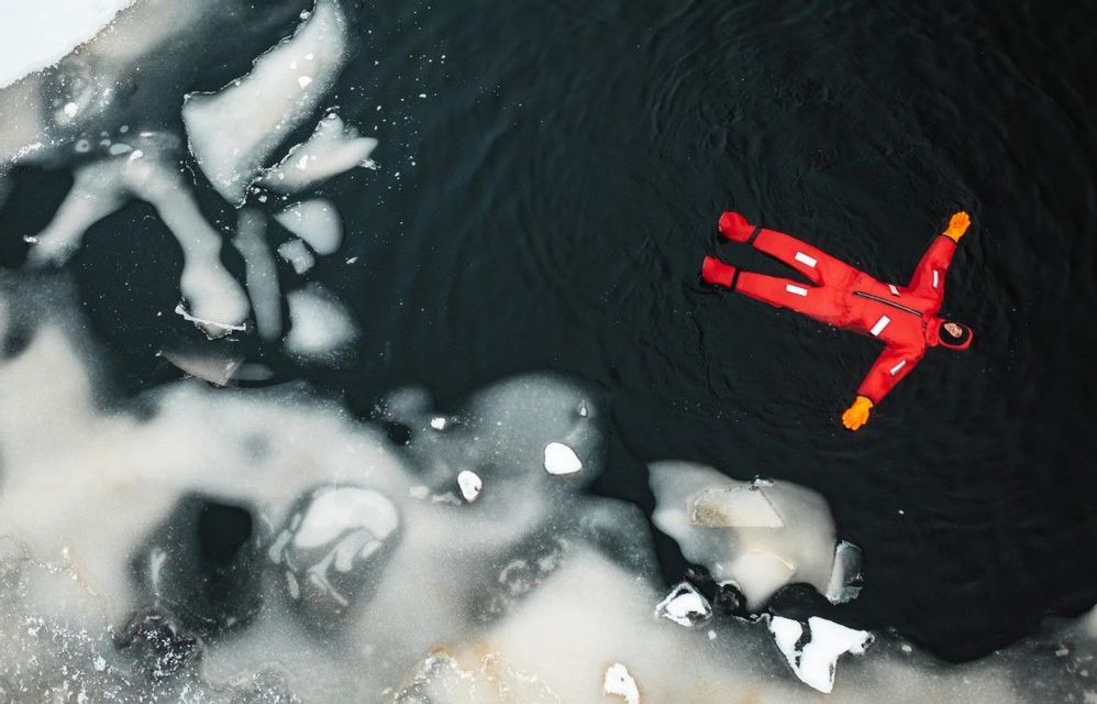 Eine Draufsicht auf eine Person in einem roten Überlebensanzug, die in dunklem, eisigem Wasser auf dem Rücken treibt.