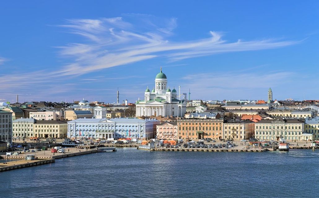 A white cathedral with a green dome stands tall over colorful waterfront buildings next to a city harbor on a clear day.