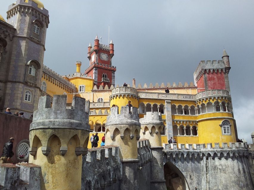Un château coloré avec des tours jaunes et rouges, vu depuis ses remparts en pierre où des gens se promènent sous un ciel nuageux.