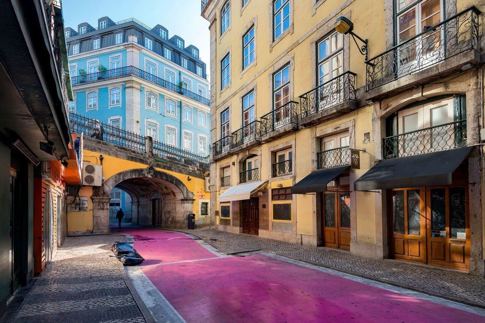 Una stretta via di città con una strada rosa brillante che passa sotto un arco in pietra, tra edifici gialli e blu.
