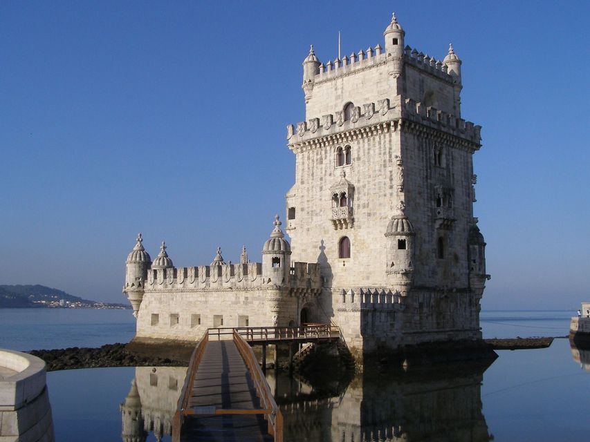 Une forteresse ornée en pierre blanche avec des tourelles est située sur des eaux calmes, accessible par un étroit pont en bois sous un ciel bleu clair.