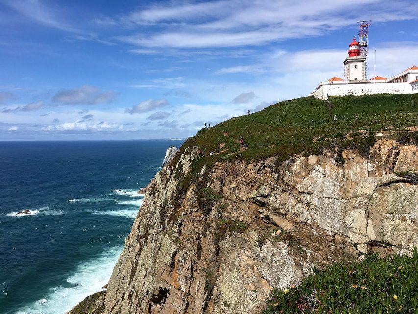 Un viaggio di gruppo WeRoad cammina su una scogliera erbosa che si affaccia sull'oceano, con un faro bianco e rosso visibile sulla cima della collina.