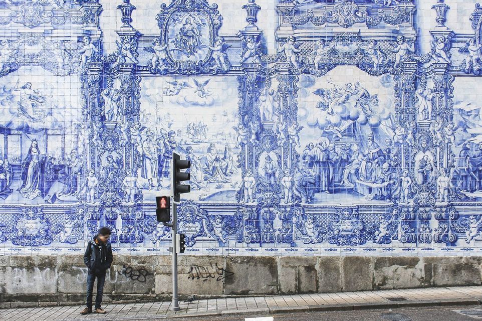 Una persona è in piedi su un marciapiede, accanto a un semaforo, davanti a un grande muro ricoperto di intricate piastrelle blu e bianche.