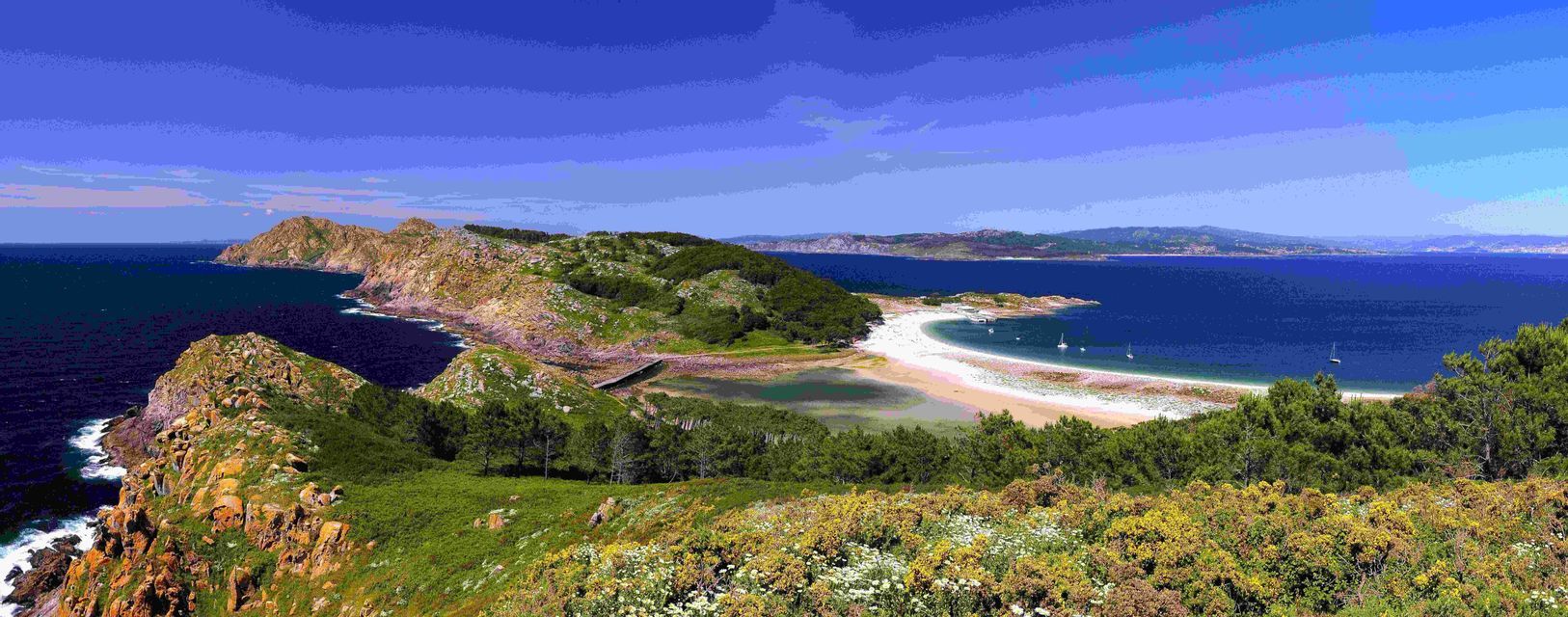 Vista panoramica di una costa verde e rocciosa con una spiaggia di sabbia bianca e una baia blu calma con barche a vela sotto un cielo sereno.