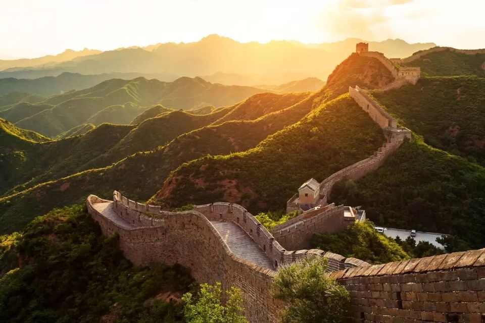 Die Chinesische Mauer schlängelt sich über sanfte grüne Berge, wobei ein Wachturm im goldenen Licht eines Sonnenuntergangs sichtbar ist.